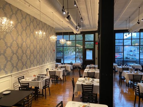 A spacious restaurant interior with elegant chandeliers, white tablecloths, and wooden floors, featuring multiple dining tables.
