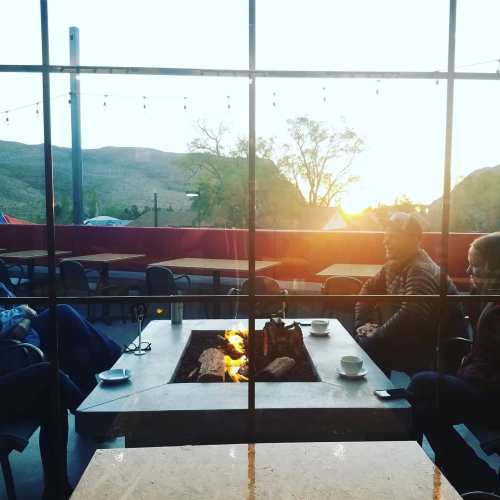 A cozy outdoor setting with a fire pit, people sitting around it, enjoying coffee as the sun sets in the background.