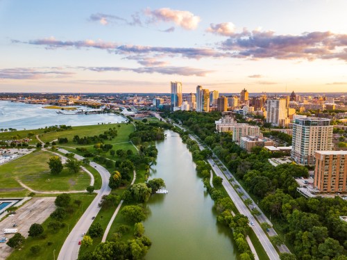 Aerial view of a city skyline at sunset, with a river and green parks in the foreground.
