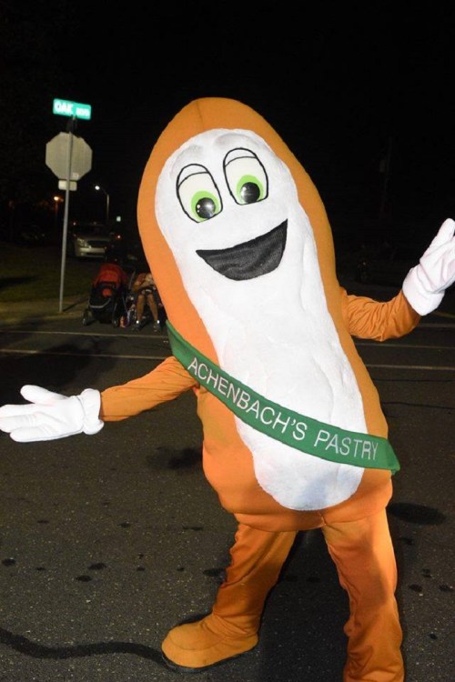 A person in a large pastry costume with a smiling face and a green sash that reads "Achenbach's Pastry."