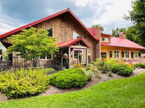 A rustic wooden building with a red roof, surrounded by greenery and flowers, features a "Welcome" sign at the entrance.