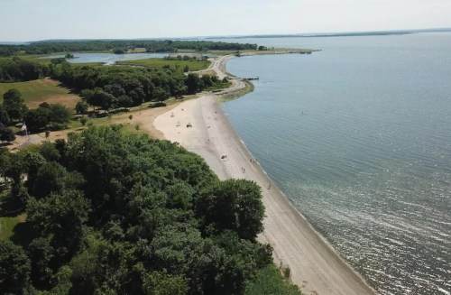Aerial view of a sandy beach along a calm shoreline, surrounded by greenery and water under a clear sky.