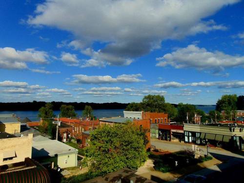 A scenic view of a small town by a river, featuring buildings, trees, and a blue sky with fluffy clouds.
