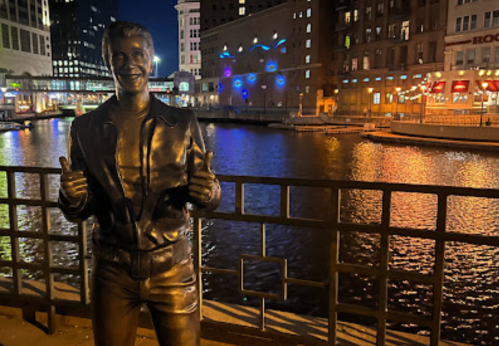 A bronze statue of a smiling man giving thumbs up, with a river and city lights in the background at night.