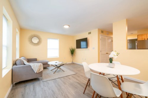 Bright living area with a gray sofa, round table, and TV, featuring yellow walls and natural light from windows.