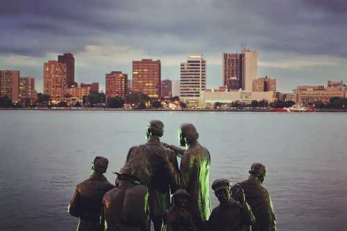 A group of bronze statues overlooking a city skyline at dusk, with buildings reflecting in the water.