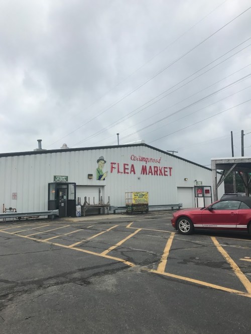 Exterior of a flea market building with a sign, cloudy sky, and a red car parked nearby.
