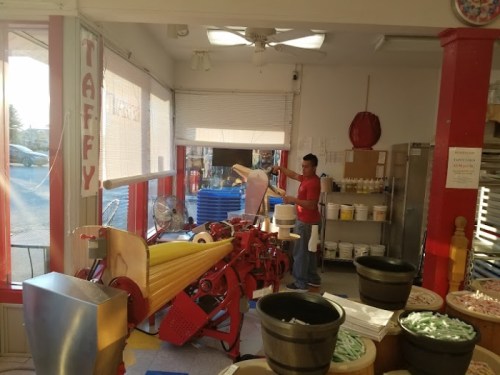 A candy shop interior with a taffy machine and a worker preparing taffy, surrounded by jars of candy and bright decor.