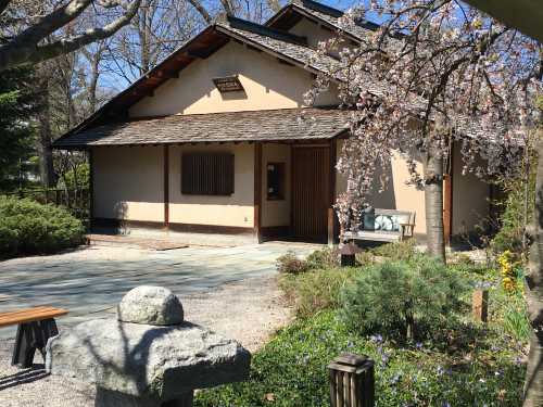 A serene garden scene featuring a traditional house, cherry blossoms, and stone pathways surrounded by greenery.