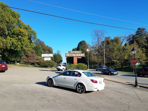 A white car parked near a sign for Kurtz Restaurant, surrounded by trees and a clear blue sky.