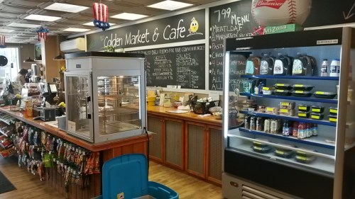 Interior of a market and café featuring a display counter, shelves of snacks, and a refrigerated drink case.