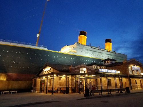 A large ship resembling the Titanic is illuminated at night, with a historic building in the foreground.