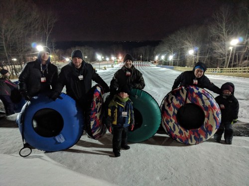 A group of five people, including a child, holding snow tubes at a nighttime tubing hill. Snowy landscape in the background.