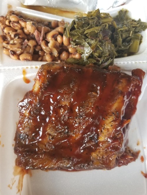 A takeout container with barbecue meat, collard greens, and black-eyed peas.