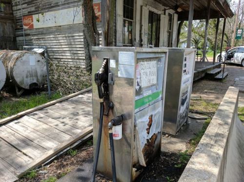 An old, rusty gas pump stands outside a weathered building, surrounded by overgrown grass and debris.
