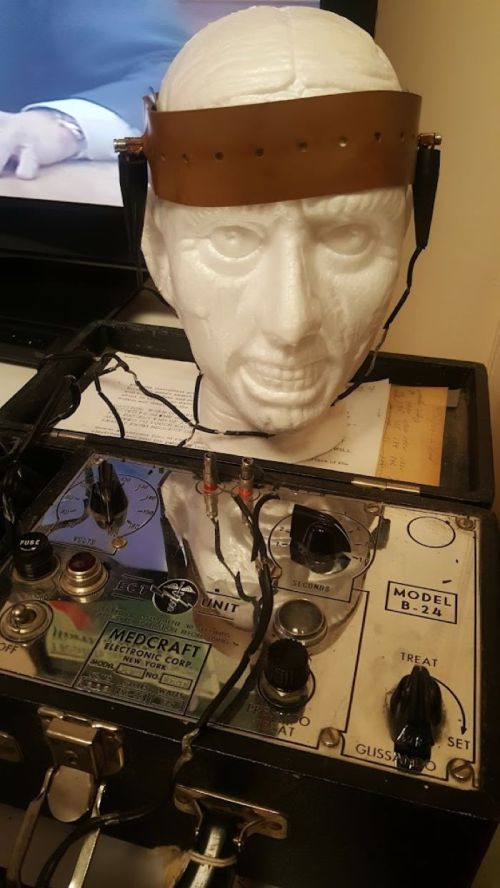 A white mannequin head with a leather strap and wires, connected to an old electronic device with dials and labels.