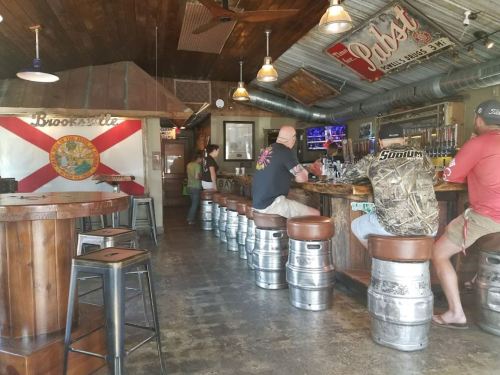 A rustic bar interior with patrons sitting on keg stools, a Florida flag on the wall, and various beer taps visible.