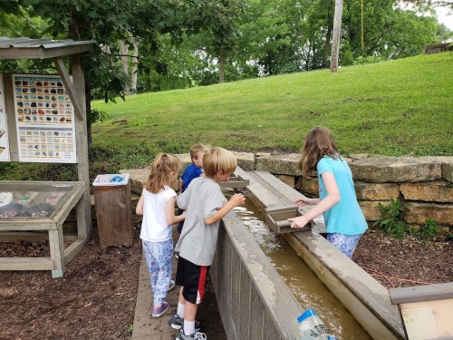 Children playing at a water table, exploring and interacting with water features in a park setting.