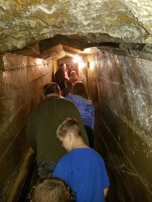 A group of people walking through a narrow, dimly lit tunnel with wet walls and a low ceiling.
