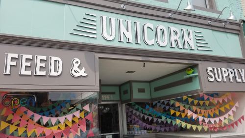 Storefront of "Unicorn Feed & Supply" with colorful bunting and an "Open" sign in the window.