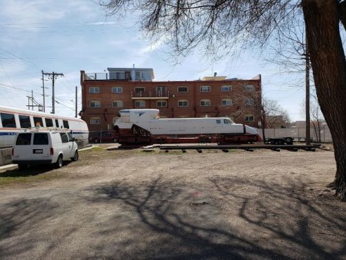 A vintage train on tracks beside a parked van and a building, with trees casting shadows in the foreground.
