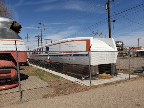A futuristic train car parked behind a fence, with power lines and a clear blue sky in the background.