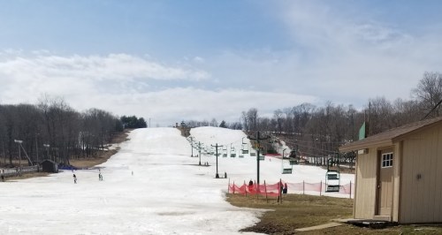 A ski slope with patches of snow, surrounded by trees, and a small building in the foreground under a cloudy sky.