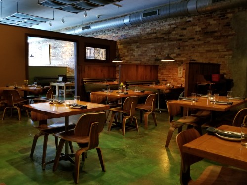 A cozy restaurant interior with wooden tables, chairs, and exposed brick walls, featuring warm lighting and a relaxed atmosphere.