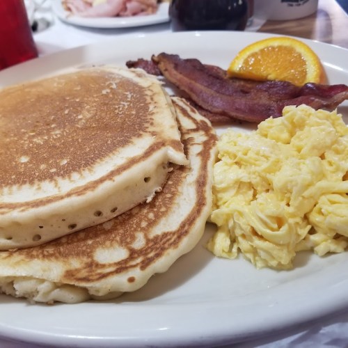 A plate of pancakes, scrambled eggs, crispy bacon, and a slice of orange.