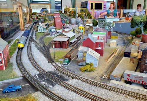 A model train layout featuring buildings, tracks, and a train, with people visible in the background.
