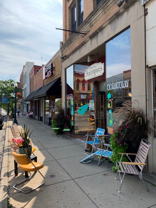 A street view featuring antique shop windows, colorful chairs, and storefronts on a sunny day.