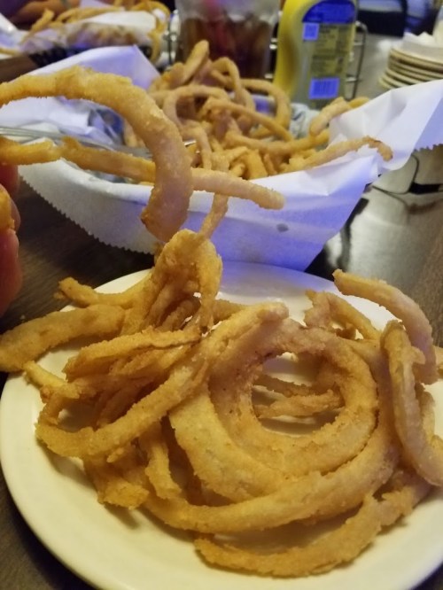 A plate of crispy, golden-brown onion rings with more in the background, served in a casual dining setting.