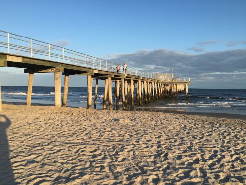 A wooden pier extends over the ocean, with people walking on it and sandy beach in the foreground under a clear sky.