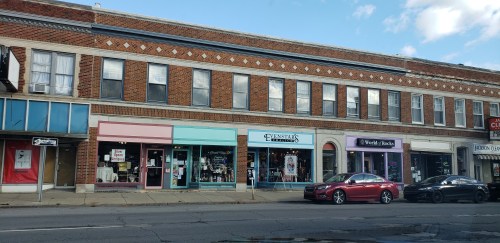 A row of storefronts featuring colorful signs, with cars parked along the street and a blue sky above.