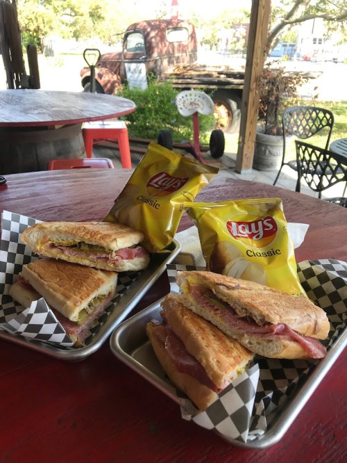 Two sandwiches on metal trays with bags of Lay's Classic chips, set on a rustic table with a vintage truck in the background.
