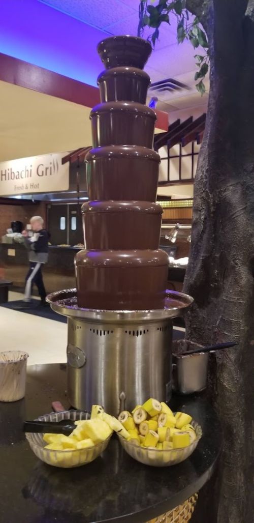 A tall chocolate fountain with bowls of sliced bananas underneath, set in a restaurant.