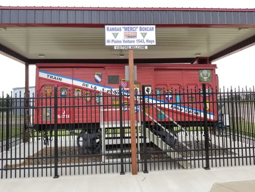 A red boxcar with colorful murals, under a shelter, surrounded by a black fence, labeled "Kansas 'MERC' Boxcar."