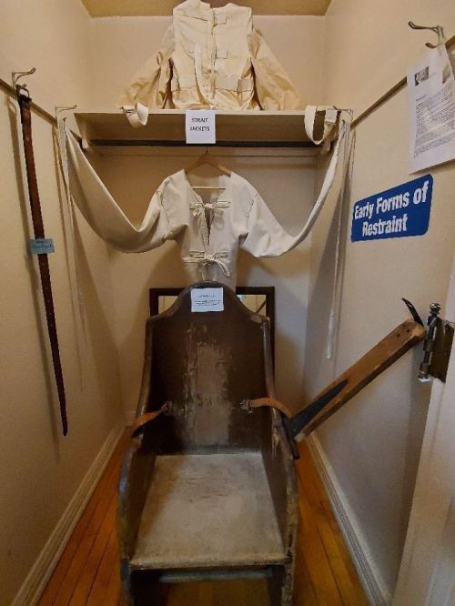 A display of early restraint devices, featuring a chair and a white restraint garment on a shelf above.