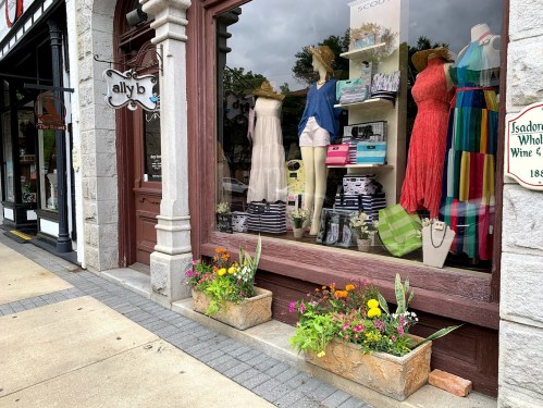A boutique window display featuring dresses, accessories, and colorful flower planters outside.