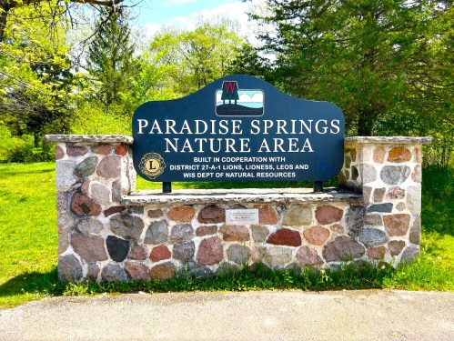 Sign for Paradise Springs Nature Area, featuring stone base and green background, surrounded by greenery.