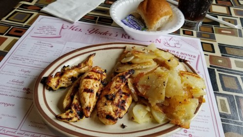 Grilled chicken strips served with seasoned fried potatoes, a roll, and a drink on a patterned table.