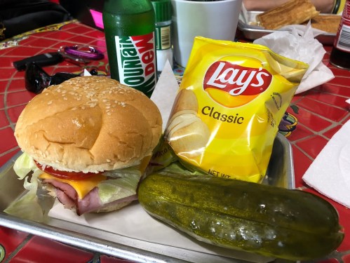 A sesame seed bun sandwich with ham, cheese, lettuce, a bag of Lay's chips, a pickle, and a bottle of Mountain Dew.