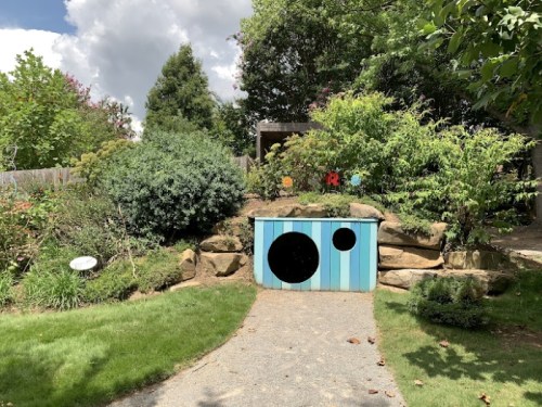 A colorful playhouse with circular cutouts, nestled in a lush garden with greenery and a gravel path.