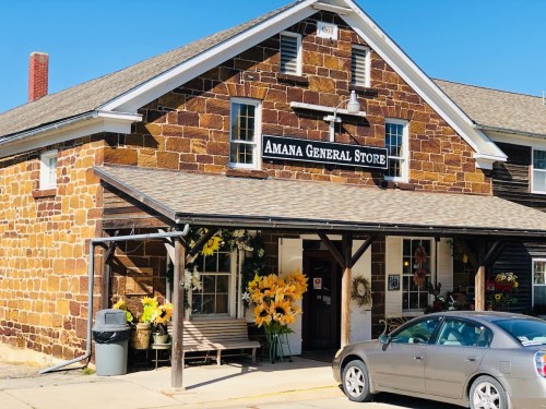 A stone building with a sign reading "Amana General Store," featuring flowers and a bench in front.