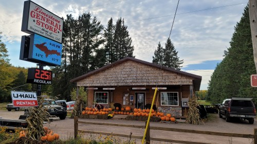 A rustic general store with a wooden exterior, pumpkins in front, and a sign displaying gas prices and U-Haul rentals.