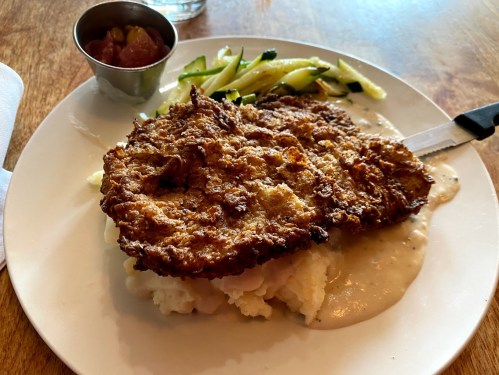 A plate with crispy fried chicken on mashed potatoes, topped with gravy, and served with sautéed zucchini and a small side of sauce.