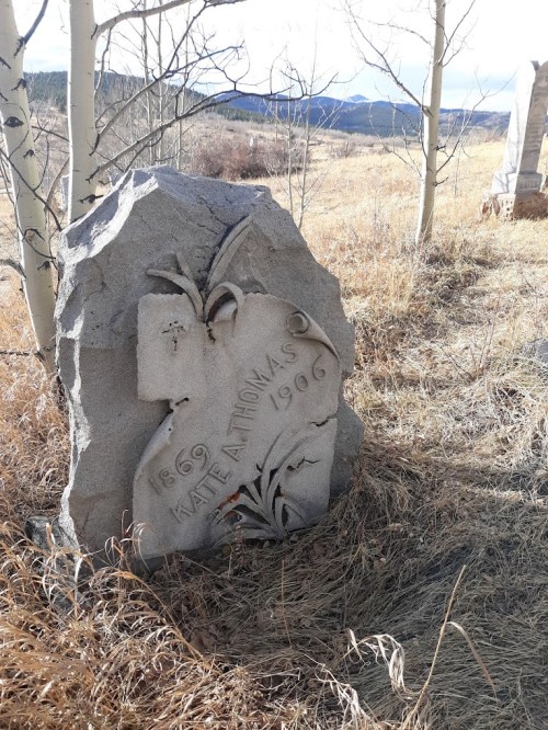 A weathered gravestone with the name "Kate A. Thomas" and dates "1869-1996," surrounded by grass and trees.