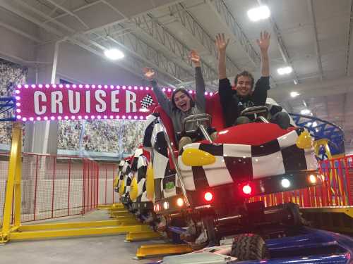 Two excited riders with hands raised on a colorful indoor roller coaster named "Cruiser."