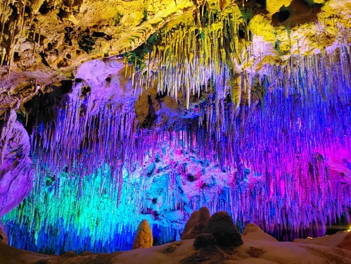 Colorful stalactites and rock formations illuminated in vibrant hues of purple, blue, and green inside a cave.
