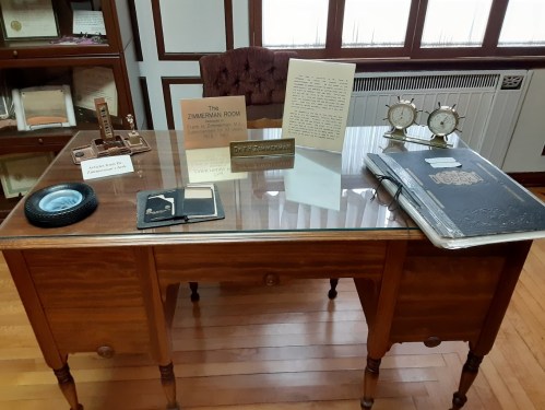 A wooden desk with a glass top, featuring a sign, a document, clocks, and various items displayed on it.
